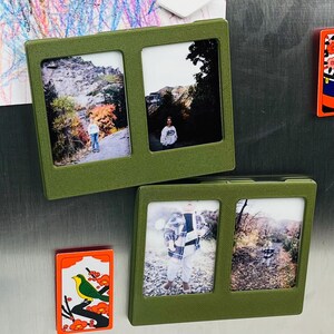 Puede incluir: Dos marcos de fotos magnéticos rectangulares de color verde oliva, cada uno con dos ranuras verticales para fotografías pequeñas. Los marcos tienen una superficie texturizada y se muestran con cuatro imágenes distintas de paisajes y retratos.