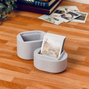 May include: Two light gray, ribbed, cylindrical containers, one holding a stack of photos. The containers are on a wooden surface, with more photos and books in the background. The photos appear to be of a landscape.