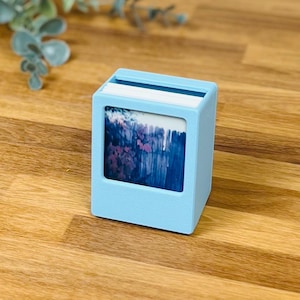 May include: A light blue square desk organizer with a matte finish, featuring a central rectangular cutout that displays a small print of a dark blue and purple floral scene. The top of the organizer has a slot containing a stack of white paper.