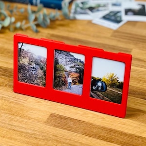 May include: A bright red rectangular triple photo frame featuring three vertical openings. The frame holds three distinct photographs: one showing a person on a path, another with a person in a rocky landscape, and a third displaying a car side mirror.