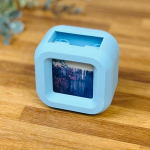 May include: A light blue cube-shaped photo frame with rounded corners and a matte finish. The frame features a central square opening displaying a printed image of a dark fence with purple flowers. The top of the frame has a rectangular slot.