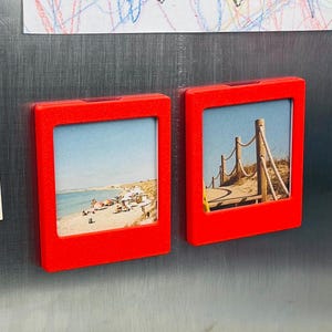 May include: Two square red plastic magnetic frames attached to a metallic surface. Each frame holds a small photograph. The left photo shows a sandy beach with umbrellas and the ocean. The right photo displays a wooden boardwalk with rope railings.