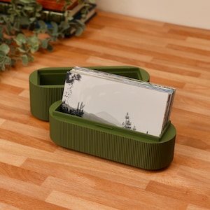 May include: An olive green oval storage box with a ribbed exterior texture, featuring an open lid and a base filled with a stack of rectangular cards. The cards display a black and white landscape illustration featuring trees and distant mountains.