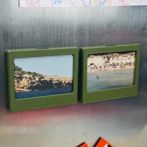 May include: Two rectangular olive green magnetic photo frames with a textured finish. The left frame displays a rocky coastal landscape, while the right frame shows a sandy beach scene with people near the water. Both frames are attached to a surface.
