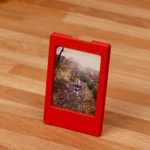 May include: A rectangular red plastic picture frame with a textured finish. The frame holds a vertical photograph depicting a person walking along a path through a wooded area with autumn foliage. The frame has a simple, solid border design.