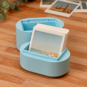 May include: A light blue oval storage container with a ribbed exterior texture, designed to hold a stack of square photographs. The container consists of a base and a removable lid, with a collection of white-bordered square prints inside.
