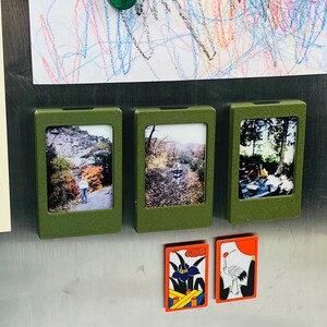 May include: Three rectangular olive green magnetic photo frames arranged horizontally, each holding a different photograph. Below them are two rectangular magnets featuring colorful graphic illustrations of a blue flower and a white crane.