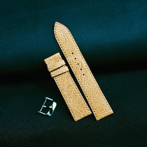 May include: Tan leather watch straps with a textured surface and a silver-colored buckle. The watch straps are shown against a dark green background. The straps are new and unused.