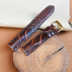 Puede incluir: Dos correas de reloj de cuero marrón con un patrón texturizado y hebillas doradas. Una correa es más larga que la otra. Las correas se muestran sobre una superficie de madera y una tela beige.