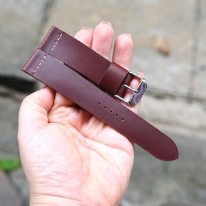 May include: A burgundy leather watch strap with a silver buckle. The strap is held in a hand and features white stitching and several holes for adjustment. The leather appears smooth and the overall design is simple and classic.