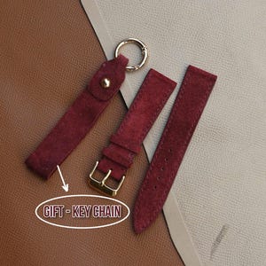 Puede incluir: Un llavero de ante burdeos con un anillo dorado y una correa de reloj a juego. El llavero tiene un cierre de botón a presión. La correa del reloj tiene una hebilla dorada. El texto "GIFT - KEY CHAIN" está en un óvalo.