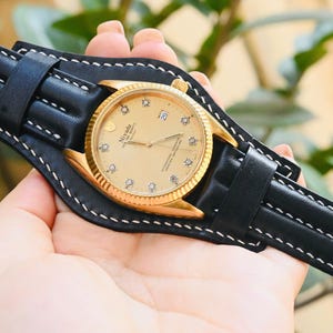Op de afbeelding: Een goudkleurig polshorloge met een lichtgekleurde wijzerplaat en diamant uurmarkeringen, gezet in een zwarte leren manchet met witte stiksels. De horlogeband is ook van zwart leer. Het horloge is een Nivada Grenchen.