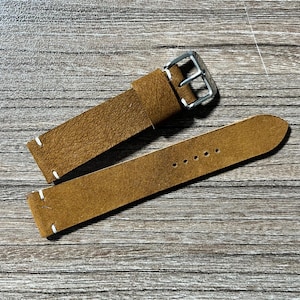 Op de afbeelding: Een bruine leren horlogeband met een zilveren gesp en witte stiksels. De band is gemaakt van een textuurrijk materiaal en heeft verschillende gaten voor aanpassing. De horlogeband wordt getoond op een houten oppervlak.