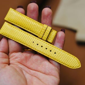 Puede incluir: Correa de reloj amarilla brillante con un patrón texturizado. La correa tiene múltiples orificios para ajuste y una hebilla. La correa del reloj se sostiene en una mano, mostrando su color y diseño.