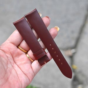 Pode incluir: Uma bracelete de relógio em pele cor de bordeaux escuro com superfície lisa, composta por duas peças. A peça mais longa tem cinco orifícios perfurados para ajuste, enquanto a mais curta inclui uma presilha fixa e um entalhe na extremidade.