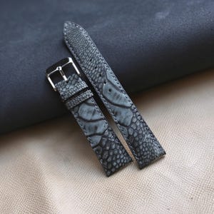 May include: Gray watch strap with a textured, patterned surface. The strap features a silver-colored buckle and a contrasting lighter gray pattern. The watch strap is made of a durable material and is designed for a watch.