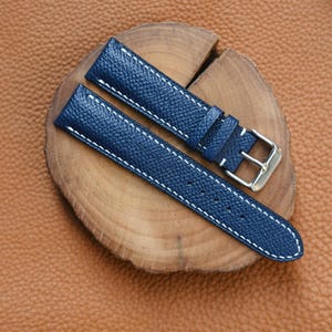 Könnte beinhalten: Ein marineblaues Uhrenarmband mit weißen Nähten und einer silbernen Schnalle. Das Lederarmband wird auf einer Holzscheibe vor einem braunen Lederhintergrund präsentiert. Das Armband ist etwa 18 cm lang.