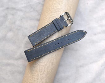 Könnte beinhalten: Zwei marineblaue Uhrenarmbänder mit gelben Nähten. Die Armbänder sind aus einem Wildleder-ähnlichen Material gefertigt und haben eine silberfarbene Schnalle. Sie liegen auf einer hellbraunen, gerollten Oberfläche.