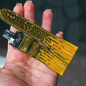 Peut inclure: Un bracelet de montre jaune vif avec un motif de peau de reptile. Le bracelet a un fermoir argenté et est tenu dans une main. Le bracelet mesure environ 20 cm de long.