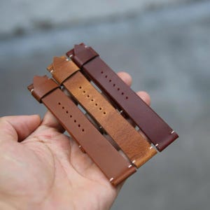 May include: Four leather watch straps in varying shades of brown, from light tan to deep burgundy. Each strap features white stitching along the edges and multiple holes for adjustment. The straps are held in a hand.