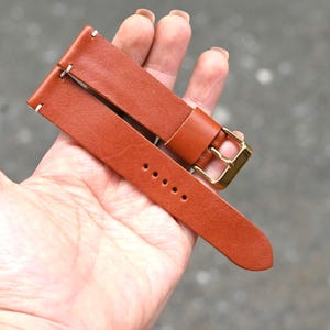 May include: A close-up of a brown leather watch strap with a gold-colored buckle. The strap has white stitching and several holes for adjustment. The leather appears to be high-quality and well-crafted.