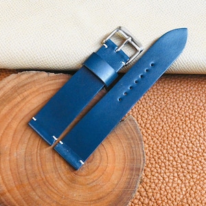 May include: A pair of blue leather watch straps with white stitching and a silver buckle. The straps are displayed on a wooden slice and a textured leather surface, showcasing the product's craftsmanship and materials.