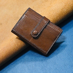 May include: A brown leather card holder with a textured, reptile-like pattern. The wallet has a flap with a button closure. The background is a mix of tan and blue leather.