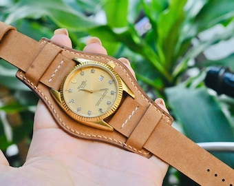 May include: A gold-toned wristwatch with a light brown leather cuff-style band. The watch face has a gold dial with diamond hour markers. The band is stitched with white thread and has a buckle closure.