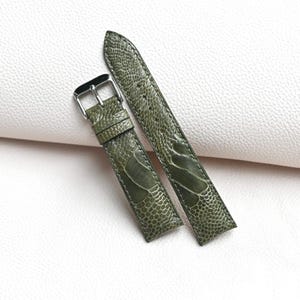 Könnte beinhalten: Zweiteiliges olivgrünes Uhrenarmband mit strukturierter, gemusterter Oberfläche. Das Armband verfügt über eine silberfarbene Schnalle und eine passende Schlaufe. Das Uhrenarmband wird auf einer weißen Oberfläche präsentiert.