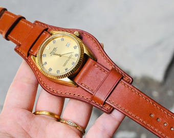 May include: A gold-toned wristwatch with a brown leather strap. The watch face has a gold finish with diamond hour markers. The watch is attached to a wide, protective leather cuff. The watch band is secured with a gold buckle.
