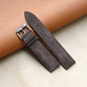 Könnte beinhalten: Zwei dunkelbraune Wildleder-Uhrenarmbänder mit einer silberfarbenen Schnalle. Die Armbänder liegen auf einer hellbraunen Lederoberfläche. Das Wildleder hat ein weiches, strukturiertes Aussehen.