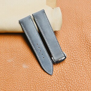 Puede incluir: Dos correas de reloj grises con costuras blancas y una hebilla plateada. Las correas están hechas de un material texturizado y se muestran sobre una superficie de cuero marrón. Las correas tienen varios agujeros para ajustar.