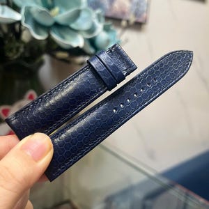 以下が含まれることがあります： 2つのパーツからなるネイビーブルーの腕時計ストラップ。六角形のテクスチャパターンが施されています。ストラップにはバックルと調整用の穴があります。時計の文字盤に取り付けるように設計されており、柔軟な素材で作られています。