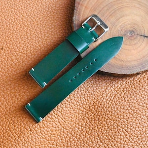 May include: A green leather watch strap with a silver buckle, lying on a textured brown leather surface. The strap is composed of two pieces, one with a buckle and the other with several holes. A wooden slice is in the background.