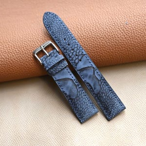 Könnte beinhalten: Zweiteiliges blaues Lederarmband mit strukturierter, gemusterter Oberfläche. Das Armband verfügt über eine silberfarbene Schnalle und wird auf einer hellbraunen und braunen Lederoberfläche präsentiert. Das Armband ist für eine Armbanduhr konzipiert.