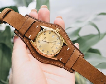 May include: A gold-toned wristwatch with a brown leather cuff band. The watch face has a gold dial with diamond hour markers. The band is a wide, wrap-around style, and the watch is held in a hand.