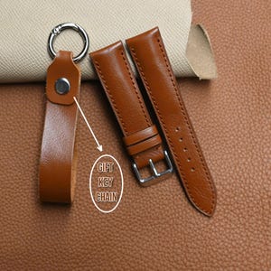 May include: A brown leather keychain with a silver ring and a pair of brown leather watch straps. The text "GIFT KEY CHAIN" is in a white oval. The items are on a brown leather surface.