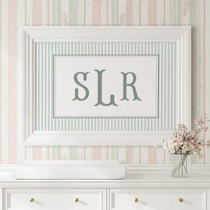May include: A white dresser with gold knobs and a changing pad sits below a framed print with the letters "SLR". The wall is decorated with vertical stripes in pink, white, and green. Two gold sconces with white shades flank the frame.