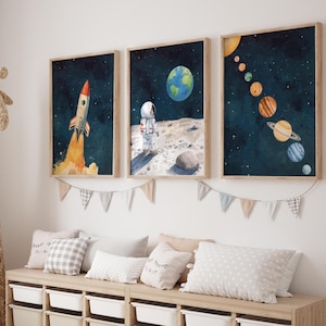 Op de afbeelding: Drie ingelijste aquarelprints met ruimtethema's. Eén toont een raket die wordt gelanceerd, een andere een astronaut op de maan met de aarde op de achtergrond en de derde de planeten in ons zonnestelsel. De prints worden boven een opbergbank weergegeven.