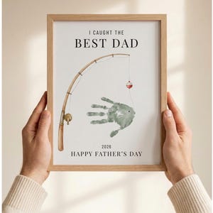 May include: A framed print with the text "I CAUGHT THE BEST DAD" above a watercolor illustration of a fishing rod and a handprint fish. The print also includes the text "HAPPY FATHER'S DAY 2026". The frame is light brown.