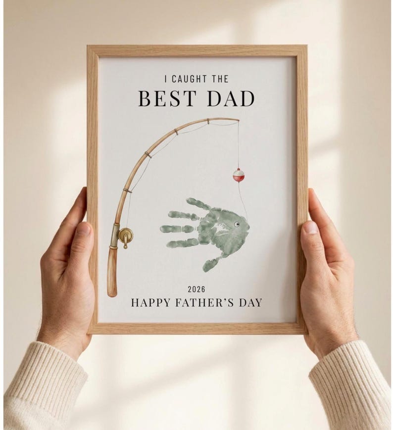 May include: A framed print with the text "I CAUGHT THE BEST DAD" above a watercolor illustration of a fishing rod and a handprint fish. The print also includes the text "HAPPY FATHER'S DAY 2026". The frame is light brown.