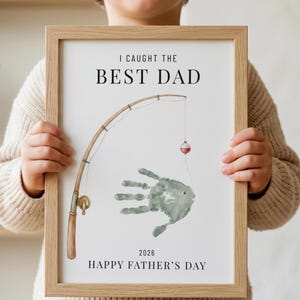 May include: A framed print with the text "I CAUGHT THE BEST DAD" and "HAPPY FATHER'S DAY". The artwork features a fishing rod, a handprint fish, and a bobber. The frame is light brown.