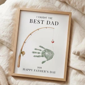 May include: Framed print with the text "I CAUGHT THE BEST DAD" above a handprint fish design and a fishing rod illustration. The bottom of the print reads "2026 HAPPY FATHER'S DAY". The frame is a light brown color.