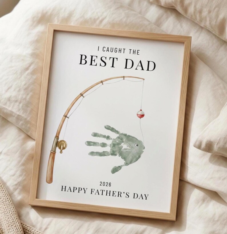 May include: Framed print with the text "I CAUGHT THE BEST DAD" above a handprint fish design and a fishing rod illustration. The bottom of the print reads "2026 HAPPY FATHER'S DAY". The frame is a light brown color.