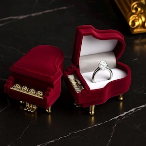 May include: Two red velvet piano-shaped ring boxes, one open to display a diamond ring. The boxes have gold-colored accents on the keys and legs. The ring features a large, clear gemstone.