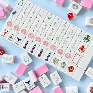 May include: A complete Mahjong set with white tiles featuring various symbols and numbers, including jokers, lighthouses, and floral designs. The set includes pink tile holders and a red die, all arranged on a light blue surface.