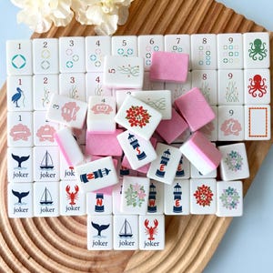 以下が含まれることがあります： 数字、花、航海をテーマにしたデザインが施された、白とピンク色の麻雀牌のセット。 木製のトレイに並べられており、ピンク色の牌が散らばっている。