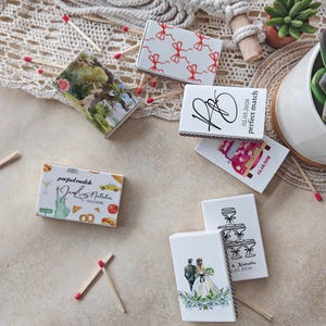 May include: Several decorative matchboxes with diverse designs are displayed. Some boxes feature illustrations of a couple, a car, and a cityscape. Other boxes have text that reads "perfect match" and dates. The matchsticks have red tips.