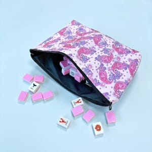 May include: A pink paisley print zippered pouch with a black interior, filled with pink and white Mahjong tiles. Some tiles are scattered outside the pouch. The tiles have various symbols and the word "Joker".