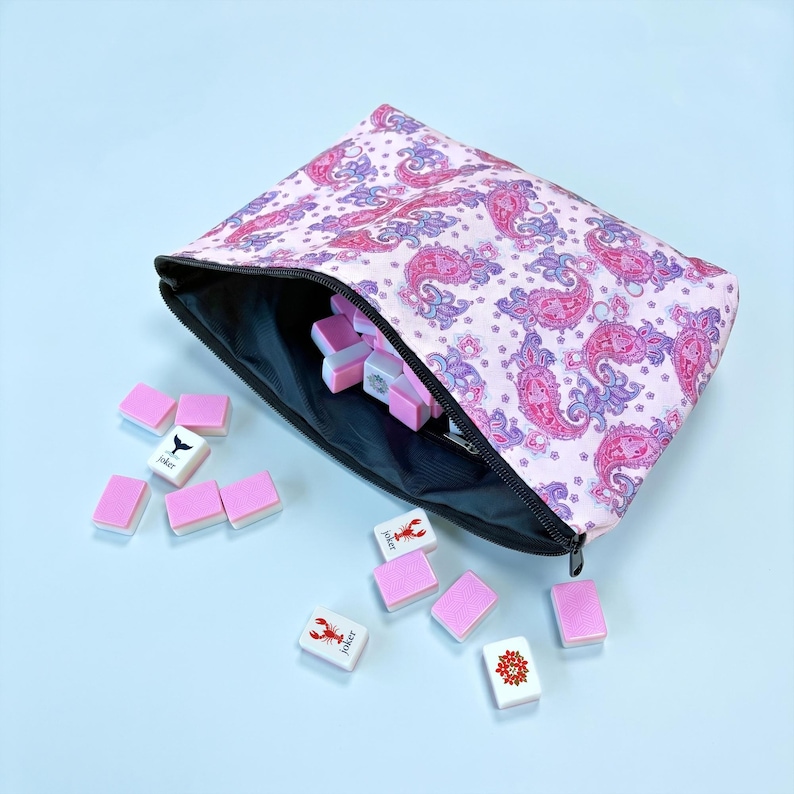 May include: A pink paisley print zippered pouch with a black interior, filled with pink and white Mahjong tiles. Some tiles are scattered outside the pouch. The tiles have various symbols and the word "Joker".